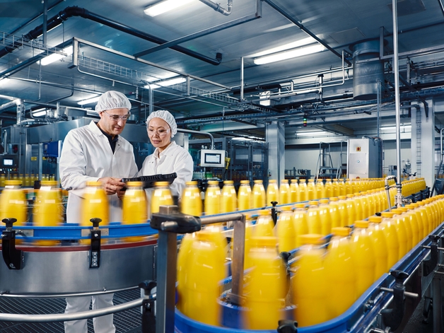 Twee technici in een drankfabriek zien toe op de botteling van sinaasappelsap.